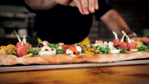 Човек прави пинса и слага подправки върху топинга й